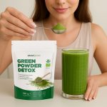 Moteruis šypsosi ir laiko šaukštelį su Detox mišinio milteliais. Detox pakuotė su stikline taip pat ant stalo